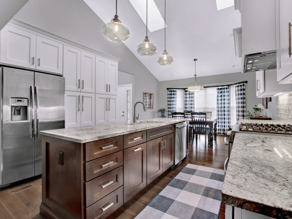 Inspiration for a mid-sized transitional medium tone wood floor, brown floor and vaulted ceiling eat-in kitchen remodel in St Louis with an undermount sink, shaker cabinets, white cabinets, granite countertops, white backsplash, subway tile backsplash, stainless steel appliances, an island and multicolored countertops