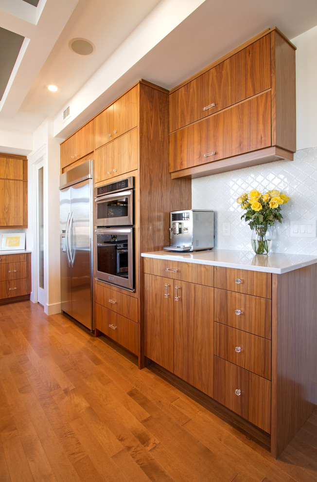 Example of a large minimalist l-shaped medium tone wood floor eat-in kitchen design with an undermount sink, flat-panel cabinets, medium tone wood cabinets, quartz countertops, white backsplash, ceramic backsplash, stainless steel appliances and an island