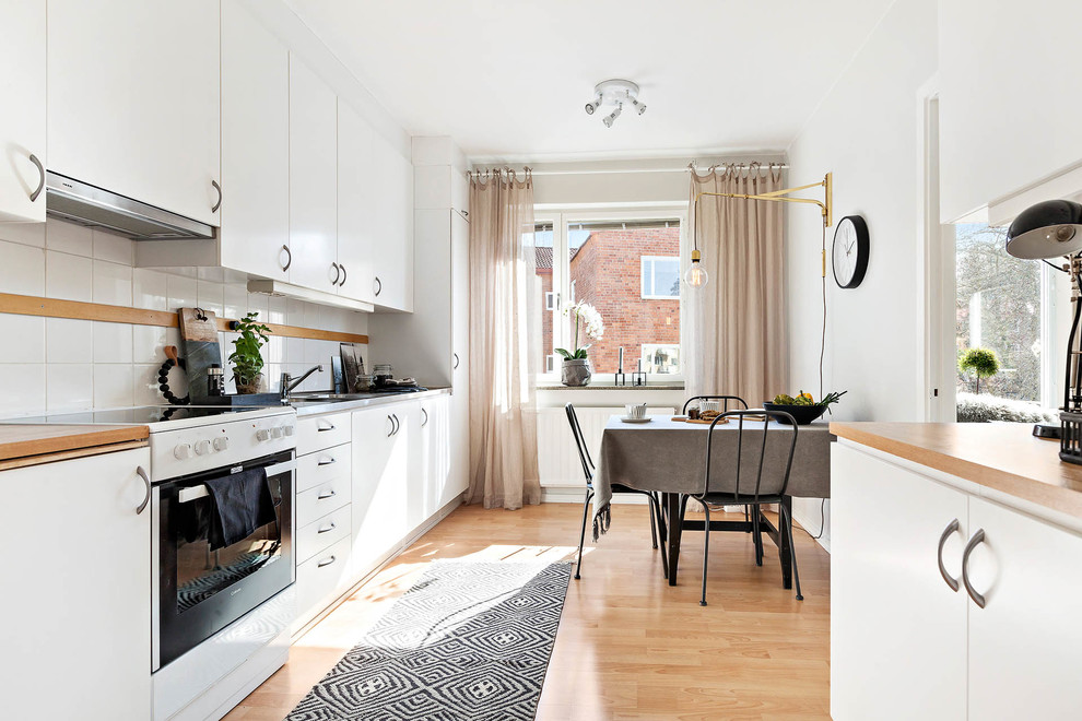 This is an example of a mid-sized scandinavian single-wall eat-in kitchen in Stockholm with flat-panel cabinets, white cabinets, wood benchtops, white splashback, white appliances, light hardwood floors and no island.