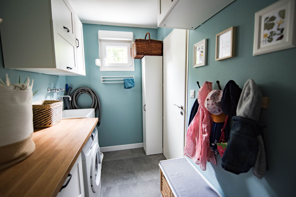Photo of a small single-wall utility room in Other with a single-bowl sink, white cabinets, wood benchtops, white splashback, cement tile splashback, blue walls, ceramic floors, an integrated washer and dryer, grey floor and yellow benchtop.