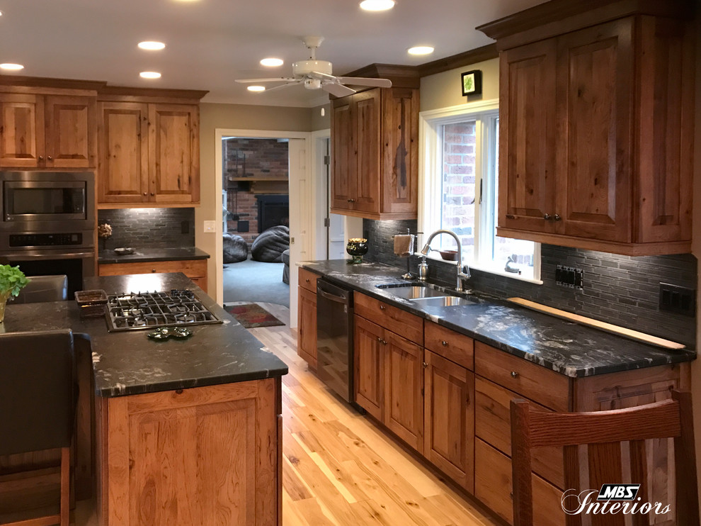 Example of a large mountain style l-shaped light wood floor and beige floor eat-in kitchen design with an undermount sink, raised-panel cabinets, medium tone wood cabinets, granite countertops, gray backsplash, ceramic backsplash, stainless steel appliances and an island