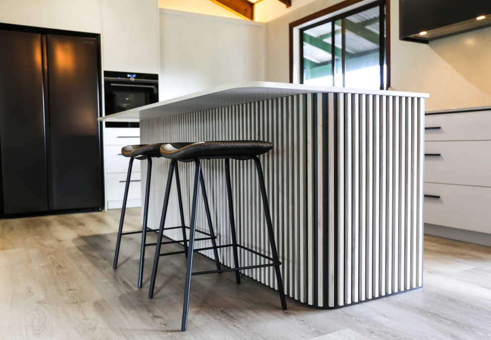 This is an example of a large contemporary l-shaped eat-in kitchen with a double-bowl sink, flat-panel cabinets, white cabinets, quartz benchtops, multi-coloured splashback, engineered quartz splashback, black appliances, laminate floors, with island, brown floor, multi-coloured benchtop and exposed beam.