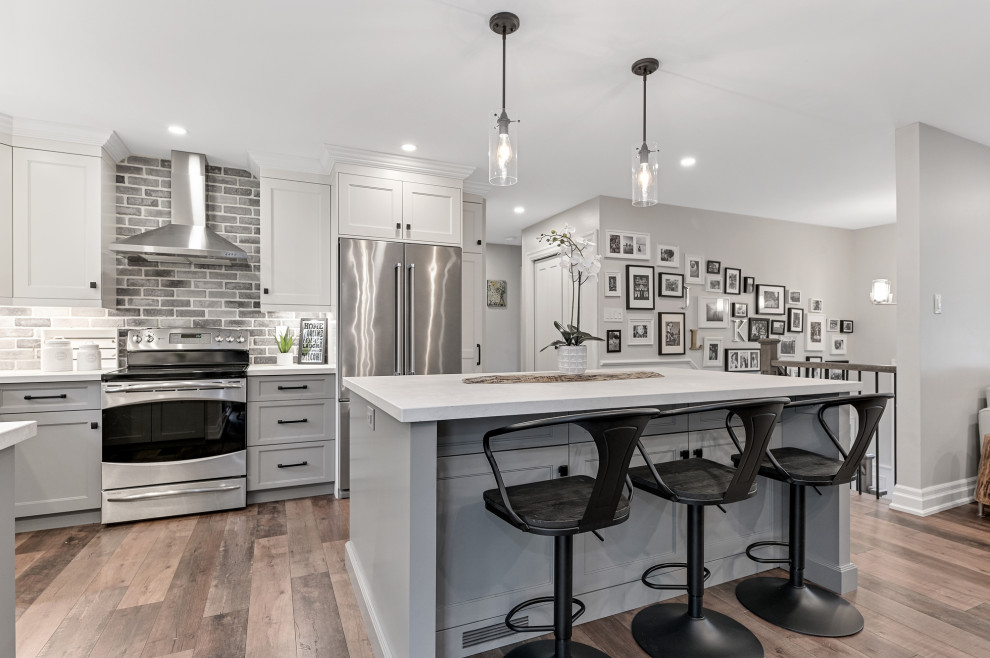 Inspiration for a mid-sized country u-shaped eat-in kitchen in Toronto with a farmhouse sink, recessed-panel cabinets, white cabinets, quartz benchtops, multi-coloured splashback, brick splashback, stainless steel appliances, laminate floors, with island, brown floor and grey benchtop.