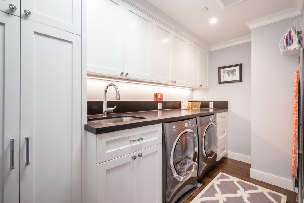 This is an example of a mid-sized transitional galley utility room in San Francisco with an undermount sink, shaker cabinets, white cabinets, quartz benchtops, grey walls, medium hardwood floors, a side-by-side washer and dryer, brown floor and grey benchtop.