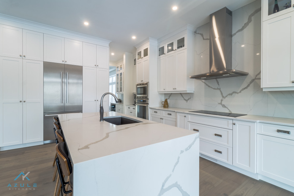Large transitional medium tone wood floor and multicolored floor kitchen photo with an undermount sink, shaker cabinets, white cabinets, quartz countertops, white backsplash, quartz backsplash, stainless steel appliances, an island and white countertops