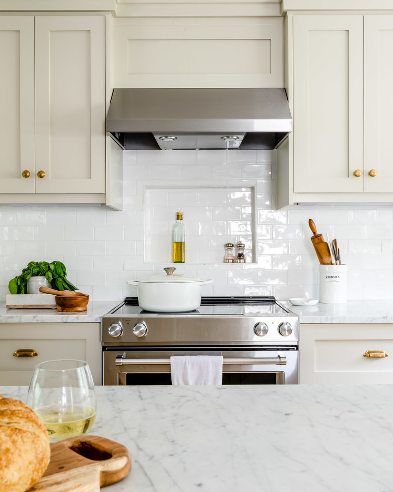 Inspiration for a mid-sized transitional l-shaped eat-in kitchen in Boston with a farmhouse sink, shaker cabinets, beige cabinets, marble benchtops, white splashback, subway tile splashback, stainless steel appliances, light hardwood floors, with island, brown floor and white benchtop.