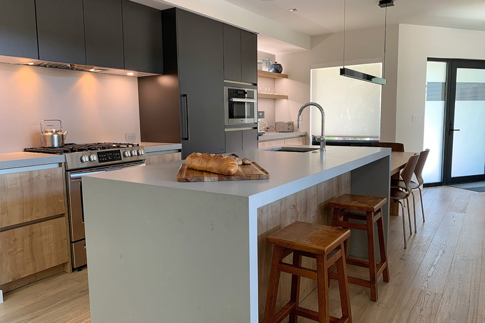 Mid-sized scandinavian l-shaped eat-in kitchen in San Francisco with an undermount sink, flat-panel cabinets, black cabinets, solid surface benchtops, grey splashback, stone slab splashback, panelled appliances, light hardwood floors, with island, brown floor and grey benchtop.