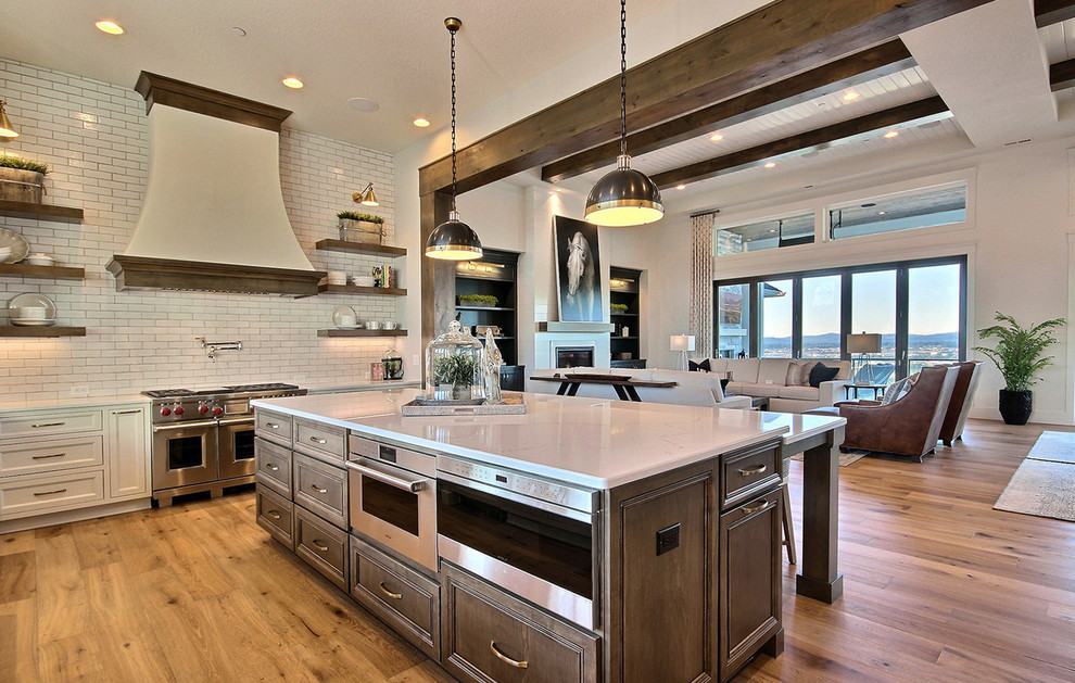 Design ideas for an expansive country u-shaped open plan kitchen in Portland with a farmhouse sink, recessed-panel cabinets, white cabinets, quartz benchtops, white splashback, ceramic splashback, stainless steel appliances, light hardwood floors, with island, brown floor and white benchtop.