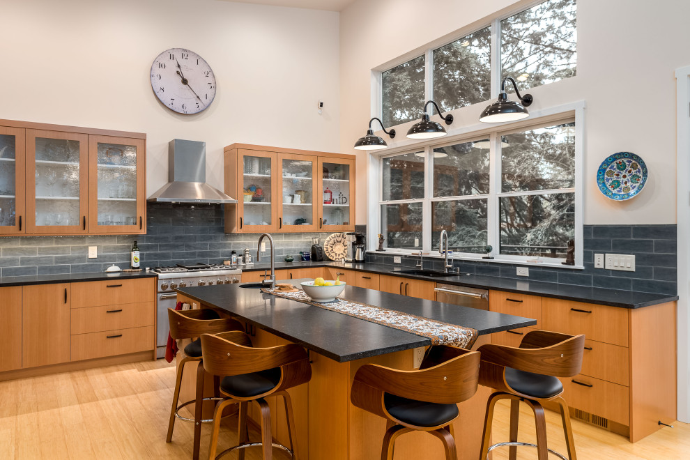 Large midcentury u-shaped eat-in kitchen in Portland with flat-panel cabinets, quartz benchtops, grey splashback, with island, black benchtop, an undermount sink, brown cabinets, glass tile splashback, stainless steel appliances, light hardwood floors and brown floor.
