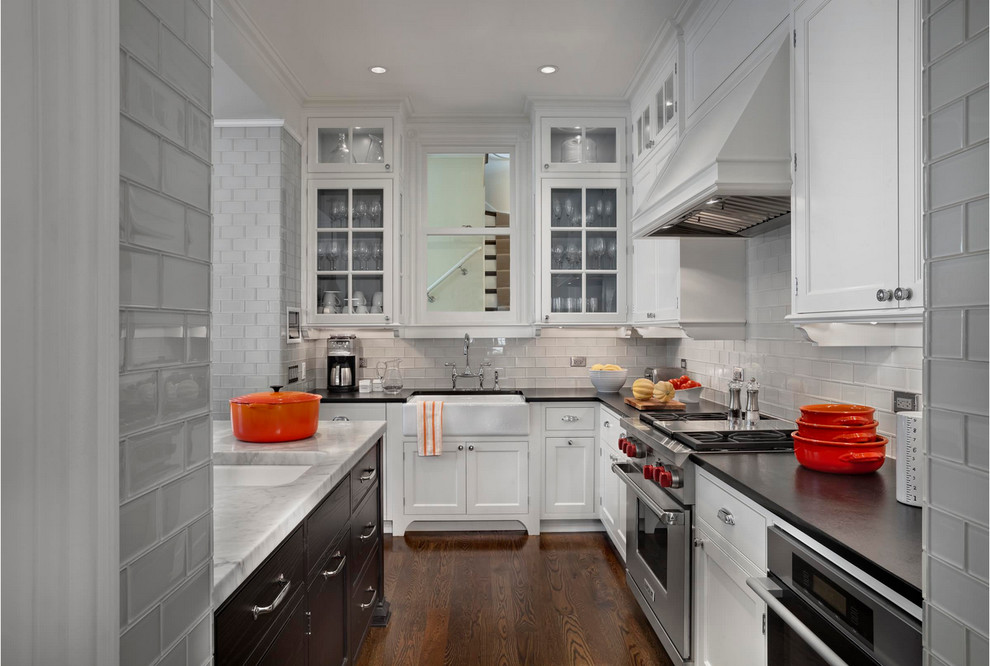 Example of a transitional dark wood floor kitchen design with a farmhouse sink, shaker cabinets, white cabinets, marble countertops, gray backsplash, glass tile backsplash, stainless steel appliances and an island
