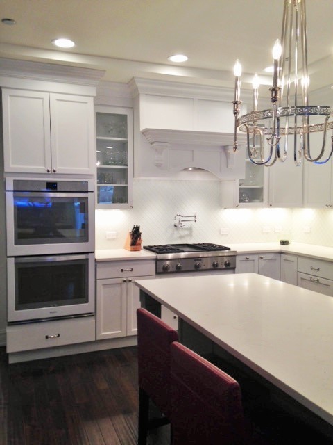 Example of a mid-sized transitional l-shaped dark wood floor eat-in kitchen design in Chicago with a farmhouse sink, recessed-panel cabinets, white cabinets, quartz countertops, white backsplash, ceramic backsplash, stainless steel appliances and an island