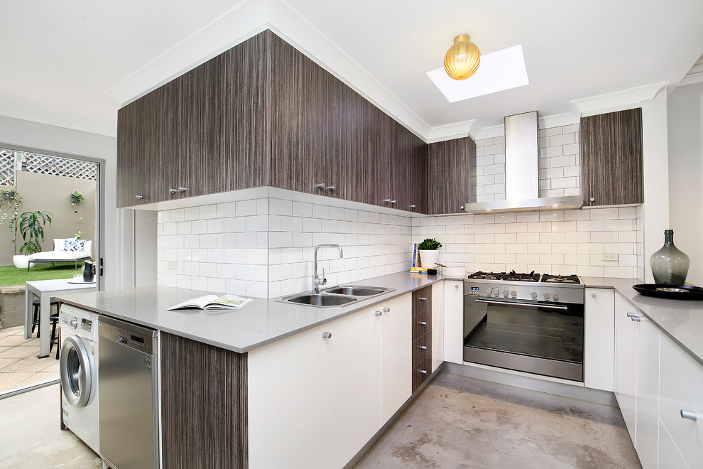 This is an example of a contemporary u-shaped kitchen in Sydney with a double-bowl sink, flat-panel cabinets, white cabinets, solid surface benchtops, white splashback, subway tile splashback, stainless steel appliances, concrete floors, a peninsula, grey benchtop and grey floor.
