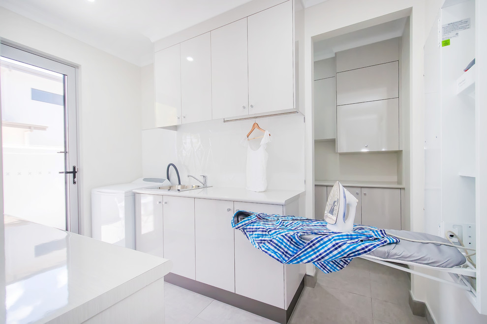 Design ideas for a large contemporary galley utility room in Other with a single-bowl sink, flat-panel cabinets, white cabinets, laminate benchtops, white walls, white splashback, glass sheet splashback, ceramic floors, a stacked washer and dryer, grey floor and white benchtop.