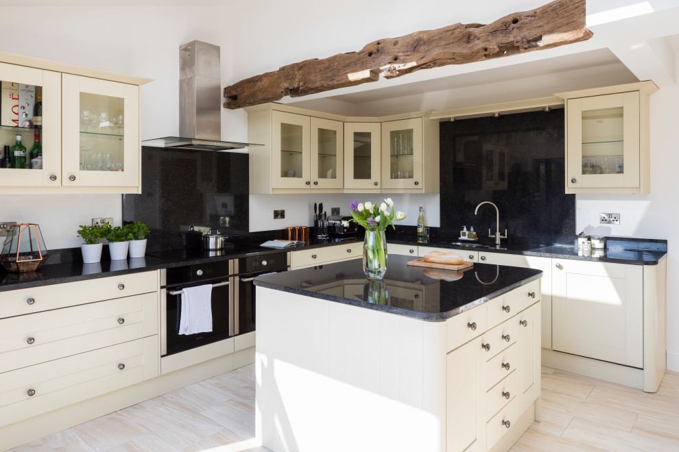 Photo of a large traditional cream and black l-shaped open plan kitchen in West Midlands with a double-bowl sink, glass-front cabinets, white cabinets, granite worktops, black splashback, laminate floors, an island, black worktops and exposed beams.