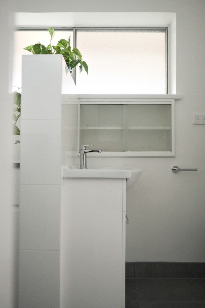 Small modern master bathroom in Melbourne with recessed-panel cabinets, white cabinets, a drop-in tub, a shower/bathtub combo, a wall-mount toilet, white tile, ceramic tile, white walls, ceramic floors, a pedestal sink, laminate benchtops, brown floor, a hinged shower door and white benchtops.