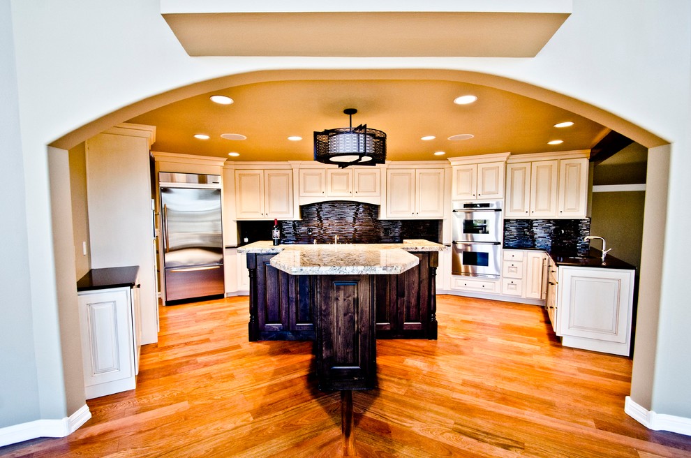 Kitchen - large transitional l-shaped medium tone wood floor and brown floor kitchen idea in Denver with white cabinets, granite countertops, black backsplash, glass tile backsplash, stainless steel appliances, an island, an undermount sink, raised-panel cabinets and multicolored countertops
