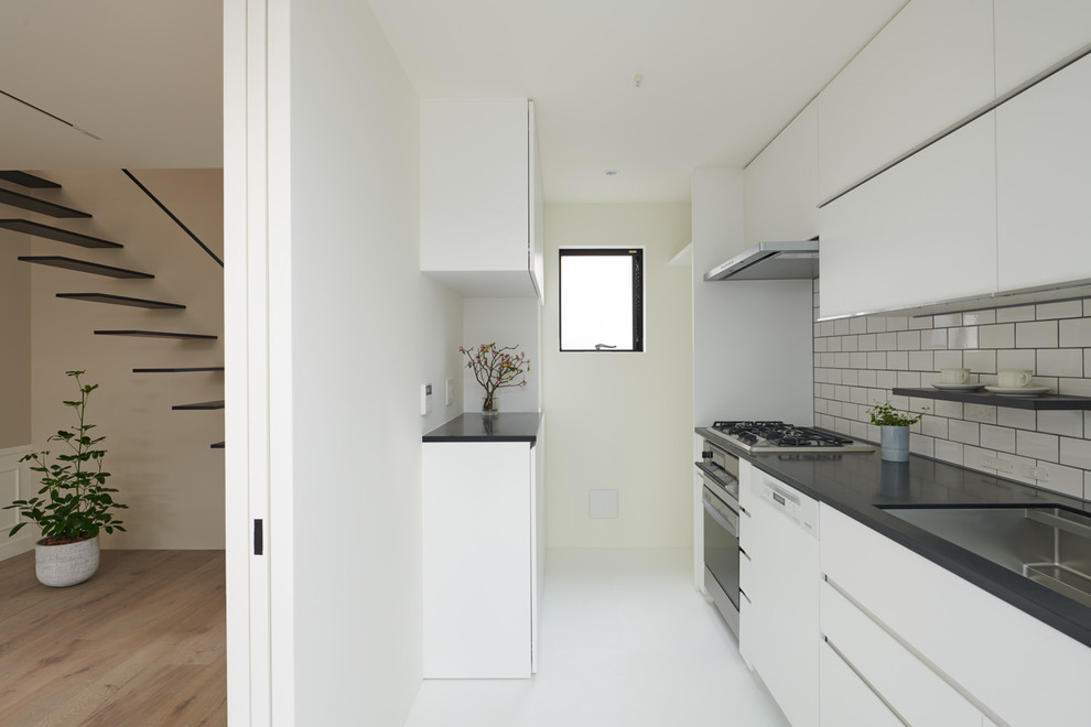 This is an example of a small modern single-wall separate kitchen in Tokyo with an undermount sink, beaded inset cabinets, white cabinets, solid surface benchtops, white splashback, timber splashback, white appliances, vinyl floors, with island, white floor and black benchtop.