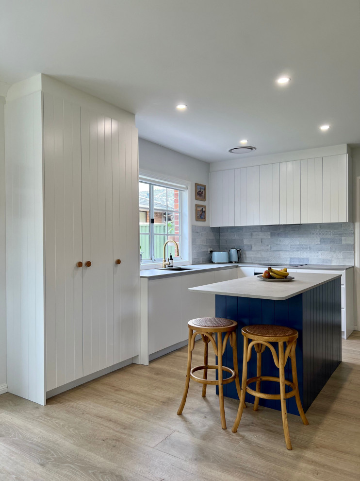 Photo of a mid-sized beach style l-shaped eat-in kitchen in Sydney with an undermount sink, recessed-panel cabinets, white cabinets, quartz benchtops, blue splashback, ceramic splashback, black appliances, light hardwood floors, with island, brown floor and grey benchtop.