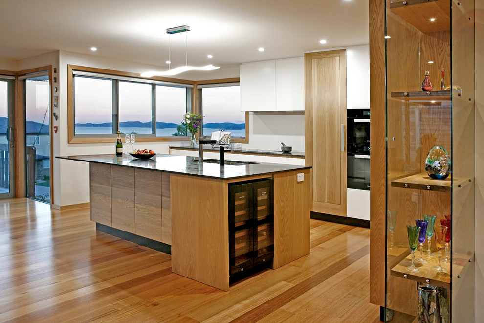 Large modern l-shaped open plan kitchen in Auckland with a double-bowl sink, recessed-panel cabinets, medium wood cabinets, granite benchtops, black appliances, medium hardwood floors, with island and black benchtop.