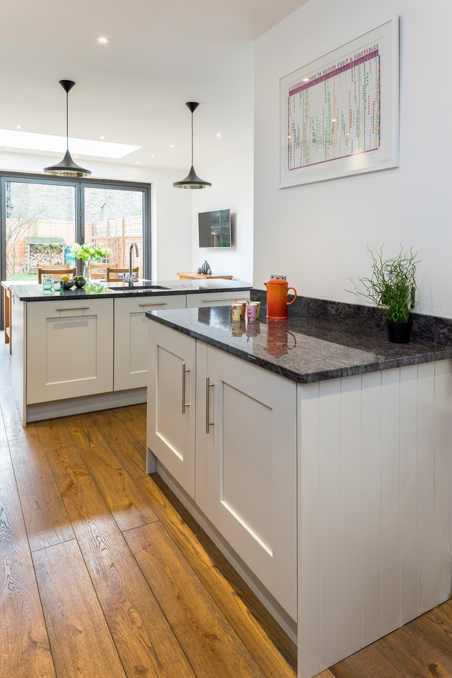 Photo of a large contemporary galley eat-in kitchen in London with an integrated sink, shaker cabinets, grey cabinets, black appliances, medium hardwood floors, with island and brown floor.