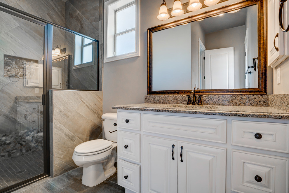 Photo of a mid-sized traditional bathroom in Austin with raised-panel cabinets, white cabinets, an alcove shower, a two-piece toilet, brown tile, gray tile, multi-coloured tile, pebble tile, ceramic floors, an undermount sink and granite benchtops.
