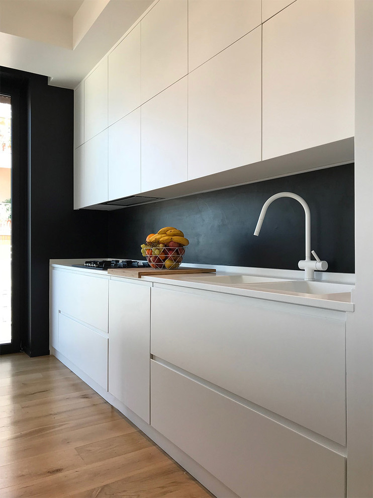 Mid-sized contemporary single-wall open plan kitchen in Rome with flat-panel cabinets, white cabinets, black splashback, an integrated sink, solid surface benchtops, stainless steel appliances, light hardwood floors, with island and white benchtop.