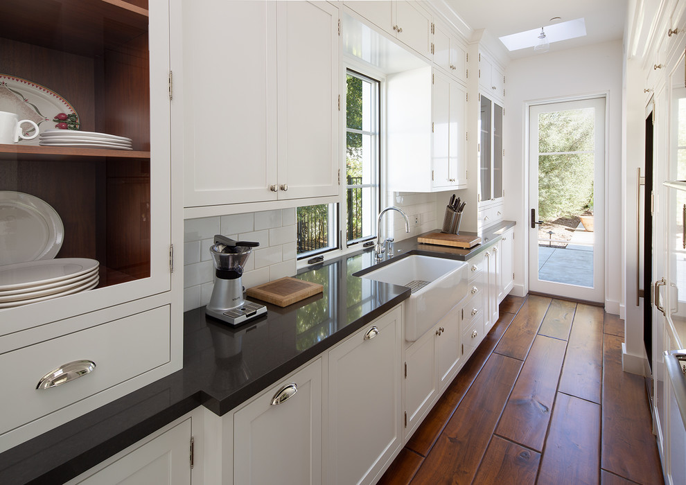 Inspiration for a mediterranean galley kitchen pantry in San Diego with a farmhouse sink, shaker cabinets, white cabinets, white splashback, subway tile splashback, medium hardwood floors, no island, brown floor and black benchtop.