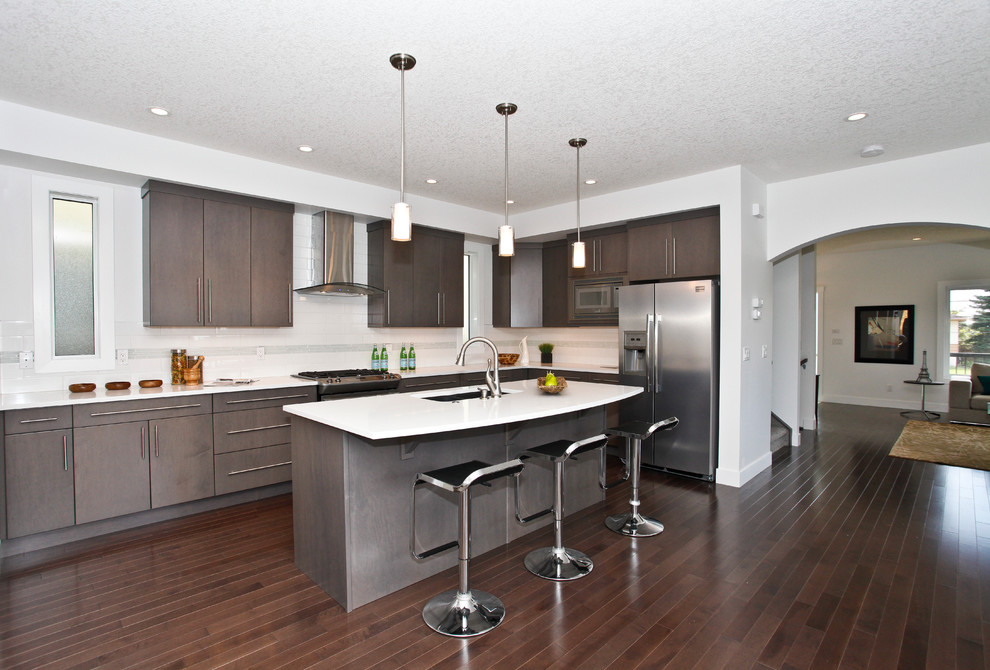 Design ideas for a mid-sized modern single-wall open plan kitchen in Calgary with stainless steel appliances, an undermount sink, flat-panel cabinets, grey cabinets, quartz benchtops, white splashback, subway tile splashback, medium hardwood floors, with island and brown floor.