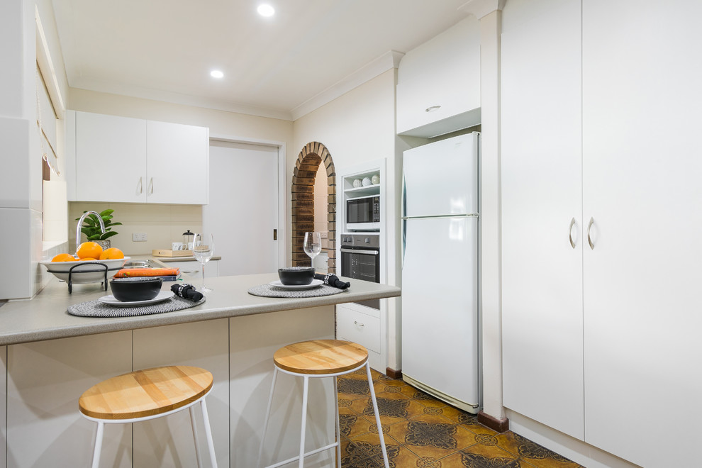 Small contemporary u-shaped kitchen in Perth with flat-panel cabinets, white cabinets, beige splashback, white appliances, a peninsula, orange floor and grey benchtop.