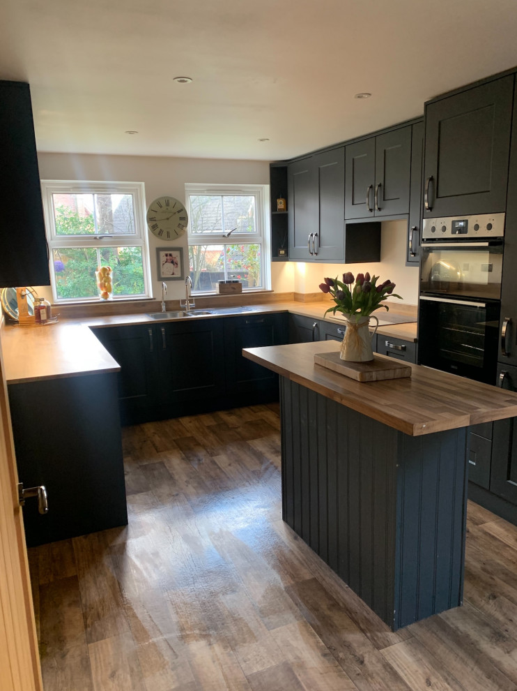 Inspiration for a medium sized traditional u-shaped enclosed kitchen in Surrey with a double-bowl sink, recessed-panel cabinets, black cabinets, laminate countertops, black appliances, medium hardwood flooring, an island, brown floors and brown worktops.