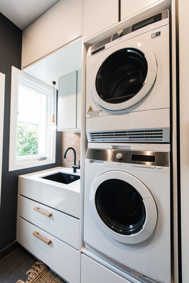 This is an example of a mid-sized contemporary single-wall dedicated laundry room in Melbourne with an undermount sink, flat-panel cabinets, white cabinets, quartz benchtops, black walls and a stacked washer and dryer.