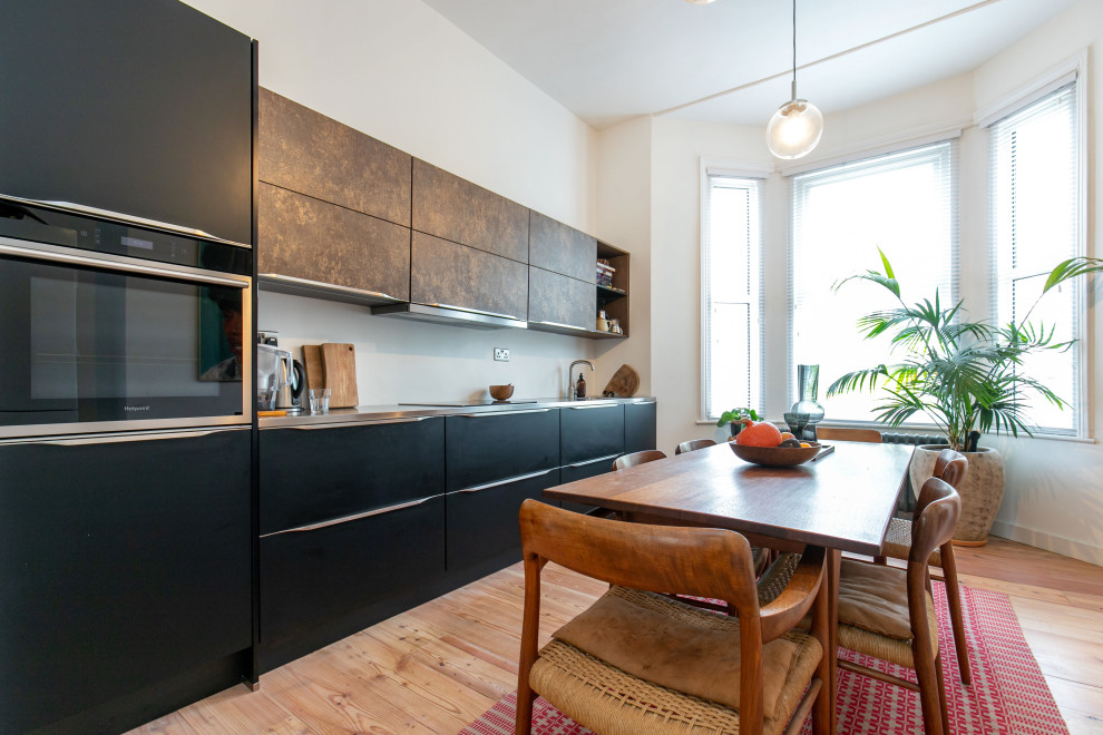 This is an example of a mid-sized modern single-wall eat-in kitchen in Sussex with an integrated sink, raised-panel cabinets, black cabinets, stainless steel benchtops, white splashback, brick splashback, black appliances, light hardwood floors, no island, beige floor, grey benchtop and coffered.