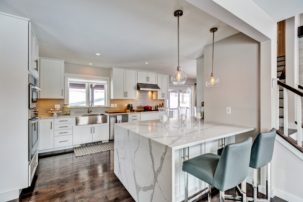 This is an example of a transitional u-shaped kitchen in Burlington with a farmhouse sink, shaker cabinets, white cabinets, beige splashback, subway tile splashback, stainless steel appliances, medium hardwood floors, a peninsula, brown floor and white benchtop.