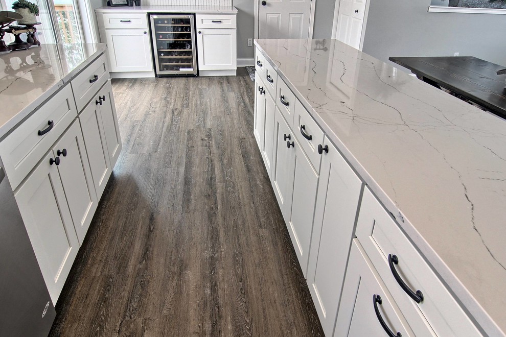 Mid-sized transitional l-shaped vinyl floor eat-in kitchen photo with a farmhouse sink, shaker cabinets, white cabinets, quartz countertops, white backsplash, stone tile backsplash, stainless steel appliances and an island