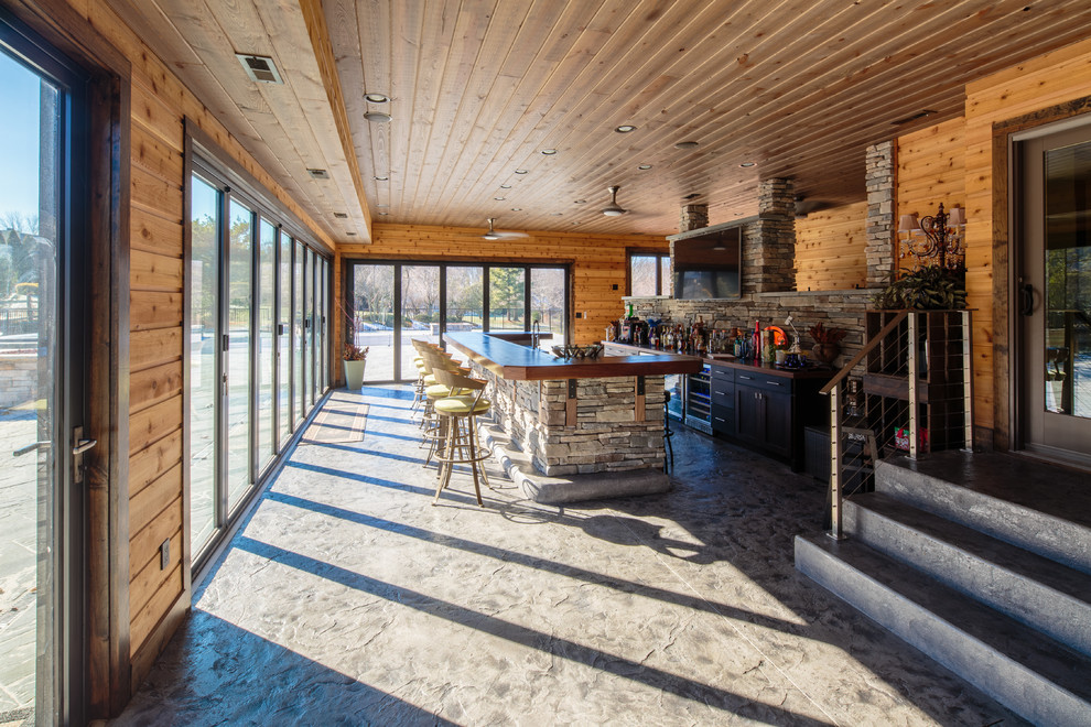 Expansive country seated home bar in Philadelphia with flat-panel cabinets, dark wood cabinets, wood benchtops, multi-coloured splashback, stone slab splashback, concrete floors and brown benchtop.