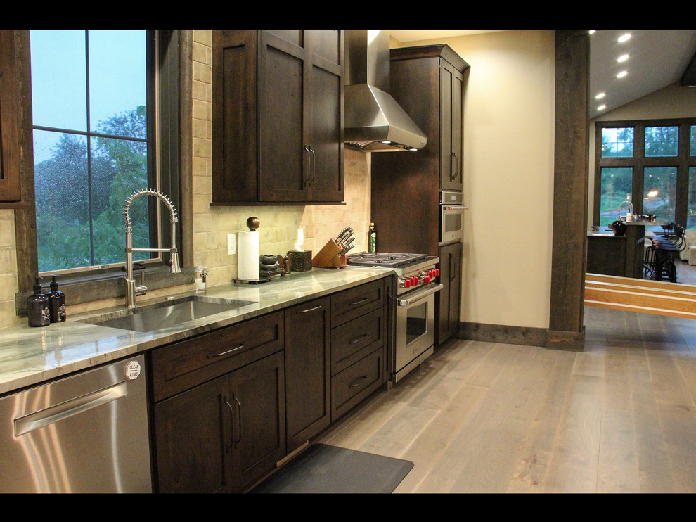 Mountain style galley medium tone wood floor and brown floor open concept kitchen photo with a single-bowl sink, shaker cabinets, dark wood cabinets, concrete countertops, gray backsplash, stainless steel appliances and gray countertops