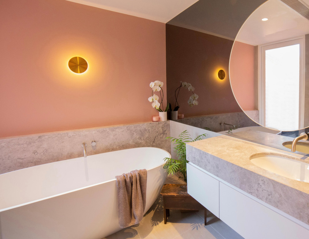 Idée de décoration pour une salle de bain de taille moyenne avec un placard à porte plane, des portes de placard blanches, une baignoire indépendante, une douche à l'italienne, un carrelage gris, des carreaux de porcelaine, un mur rose, un sol en carrelage de porcelaine, un lavabo encastré, un plan de toilette en calcaire, un sol gris, aucune cabine, un plan de toilette gris, meuble simple vasque, meuble-lavabo suspendu et un mur en pierre.