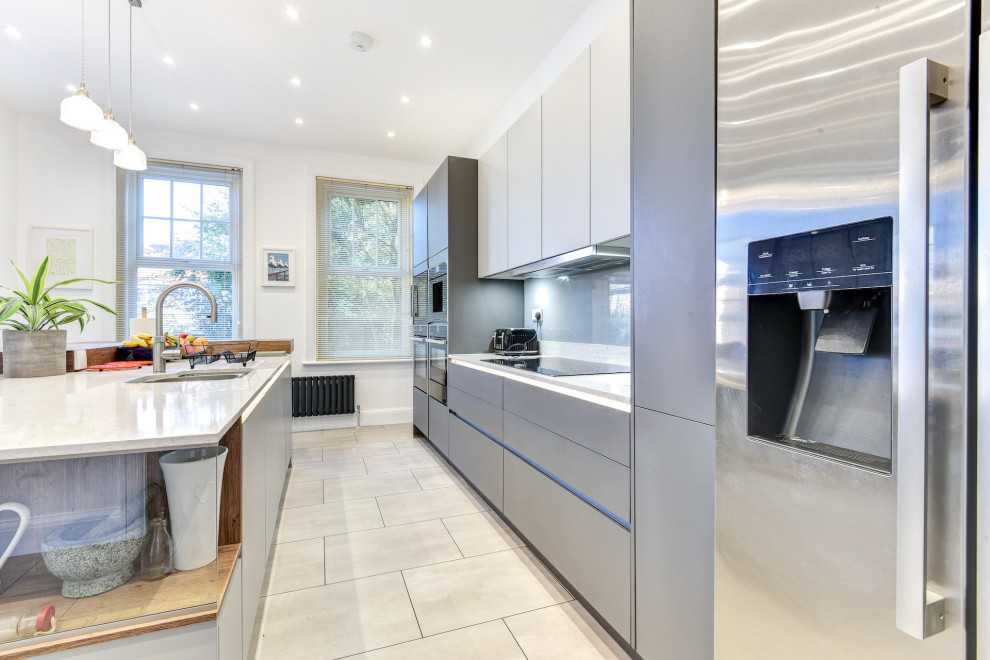 Photo of an expansive modern grey and white single-wall kitchen/diner in Sussex with a double-bowl sink, flat-panel cabinets, grey cabinets, quartz worktops, grey splashback, glass sheet splashback, black appliances, vinyl flooring, an island, grey floors, white worktops and feature lighting.