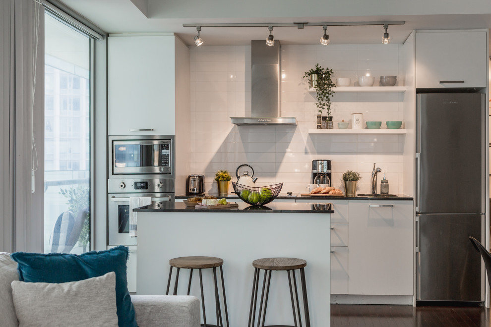 Design ideas for a contemporary single-wall open plan kitchen in Toronto with an undermount sink, flat-panel cabinets, white cabinets, white splashback, stainless steel appliances, dark hardwood floors, with island, brown floor and black benchtop.