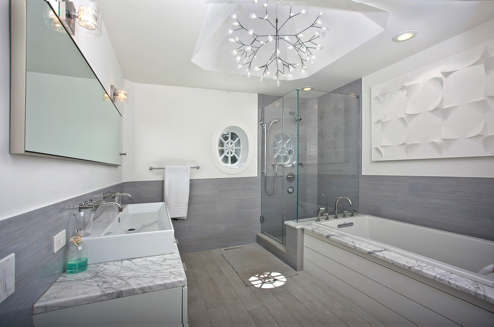 Mid-sized beach style master bathroom in New York with flat-panel cabinets, white cabinets, a drop-in tub, a corner shower, a one-piece toilet, gray tile, ceramic tile, white walls, light hardwood floors, a trough sink, marble benchtops, beige floor, a hinged shower door and white benchtops.
