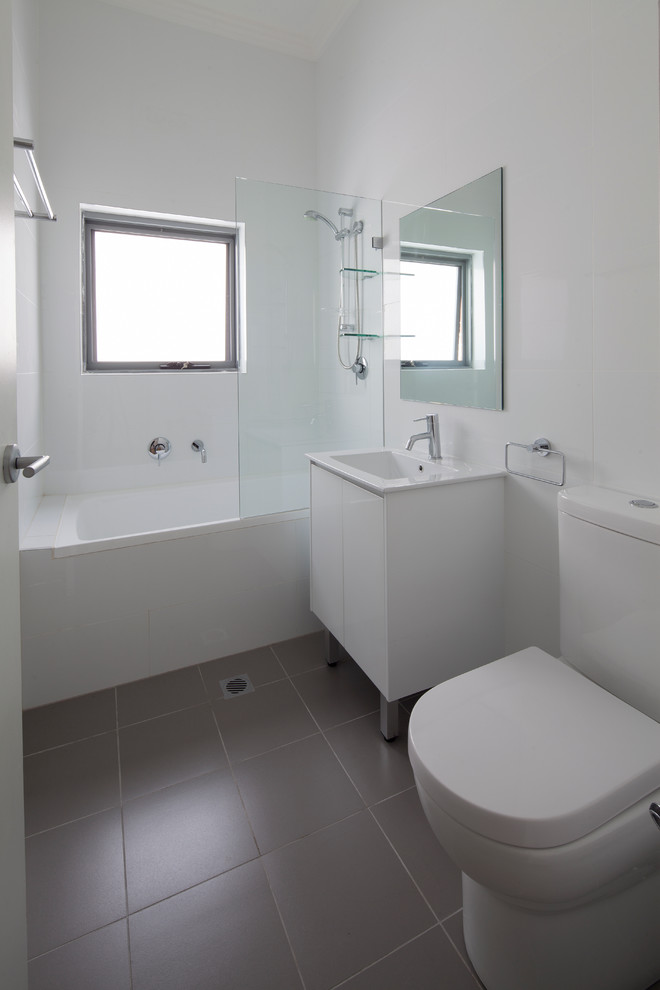 Photo of a small contemporary kids bathroom in Sydney with flat-panel cabinets, white cabinets, a drop-in tub, a shower/bathtub combo, a one-piece toilet, white tile, ceramic tile, white walls, ceramic floors, an integrated sink, grey floor, a hinged shower door and white benchtops.