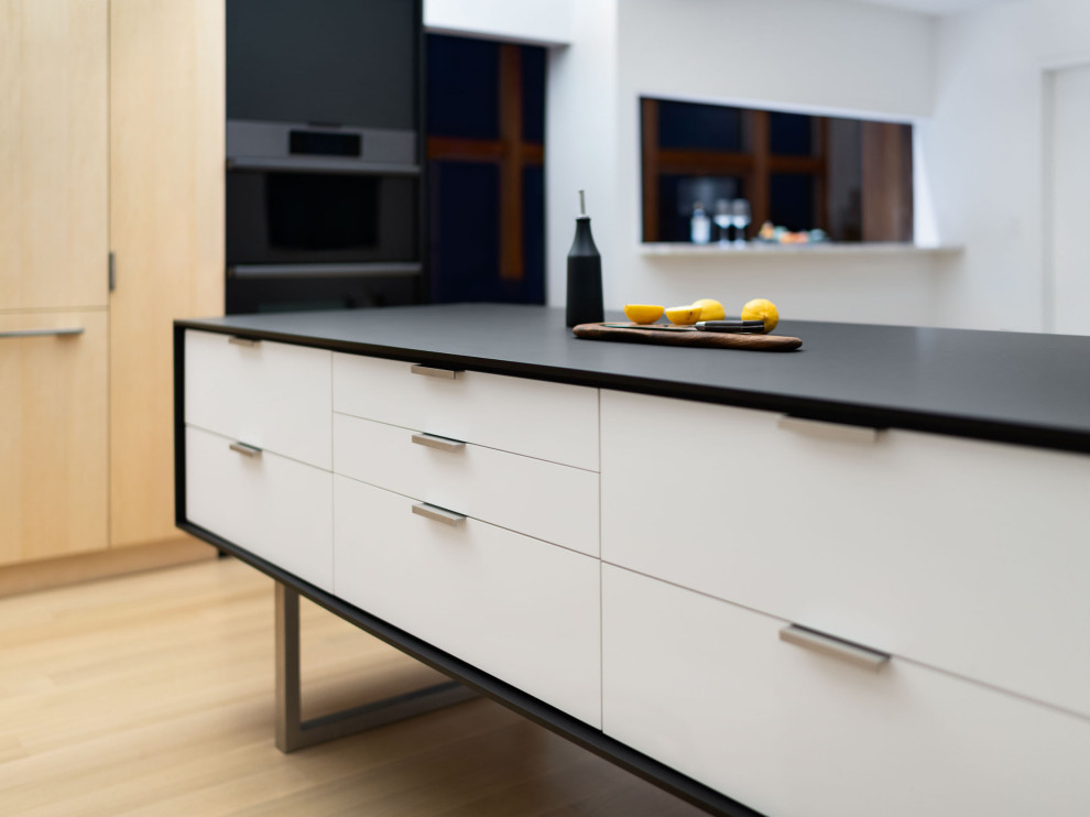 Inspiration for a modern kitchen in New York with an undermount sink, flat-panel cabinets, white cabinets, laminate benchtops, white splashback, ceramic splashback, black appliances, light hardwood floors, with island, black benchtop and vaulted.
