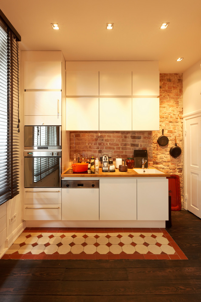 Inspiration for an expansive contemporary single-wall open plan kitchen in Hamburg with a drop-in sink, flat-panel cabinets, white cabinets, wood benchtops, red splashback, brick splashback, stainless steel appliances, dark hardwood floors, no island and brown floor.