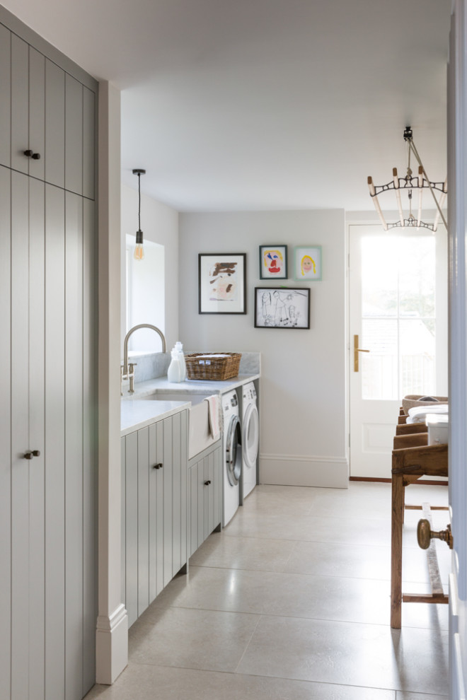 This is an example of a large transitional single-wall utility room in London with a farmhouse sink, louvered cabinets, grey cabinets, white walls, porcelain floors, a side-by-side washer and dryer, grey floor and white benchtop.
