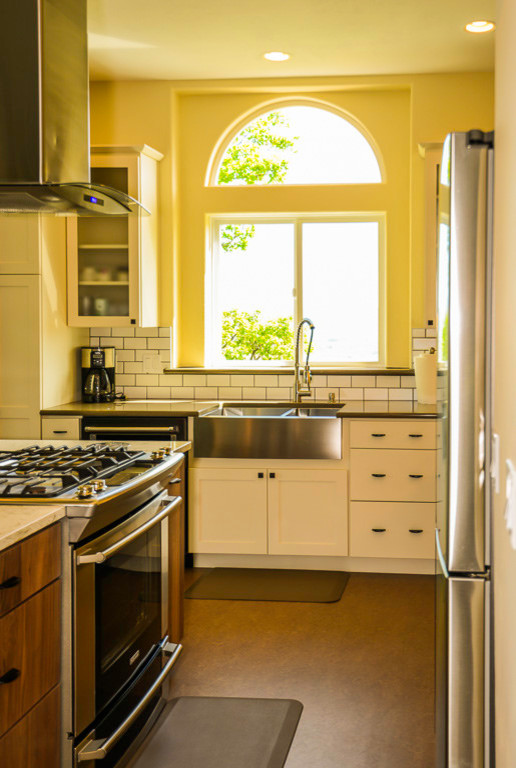 Inspiration for a mid-sized contemporary l-shaped open plan kitchen in Portland with a farmhouse sink, shaker cabinets, white cabinets, white splashback, subway tile splashback, stainless steel appliances, vinyl floors, with island and brown floor.
