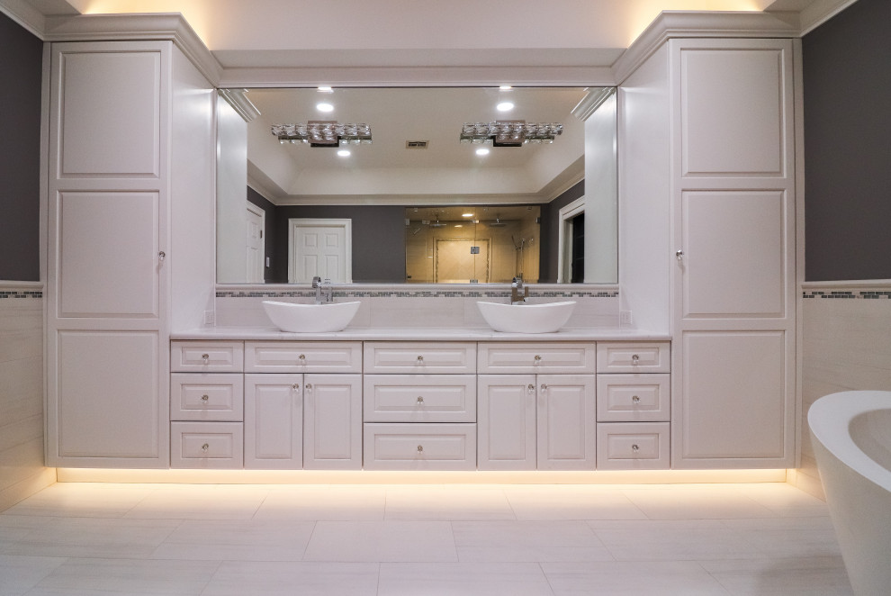Photo of an expansive transitional master bathroom in Bridgeport with raised-panel cabinets, white cabinets, a freestanding tub, a curbless shower, a one-piece toilet, white tile, porcelain tile, grey walls, porcelain floors, a vessel sink, engineered quartz benchtops, white floor, a hinged shower door and white benchtops.