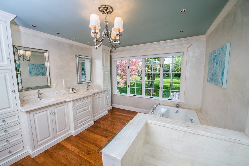 This is an example of a mid-sized traditional master bathroom in Philadelphia with an integrated sink, raised-panel cabinets, white cabinets, a drop-in tub, an open shower, a one-piece toilet, beige tile, porcelain tile, white walls and medium hardwood floors.