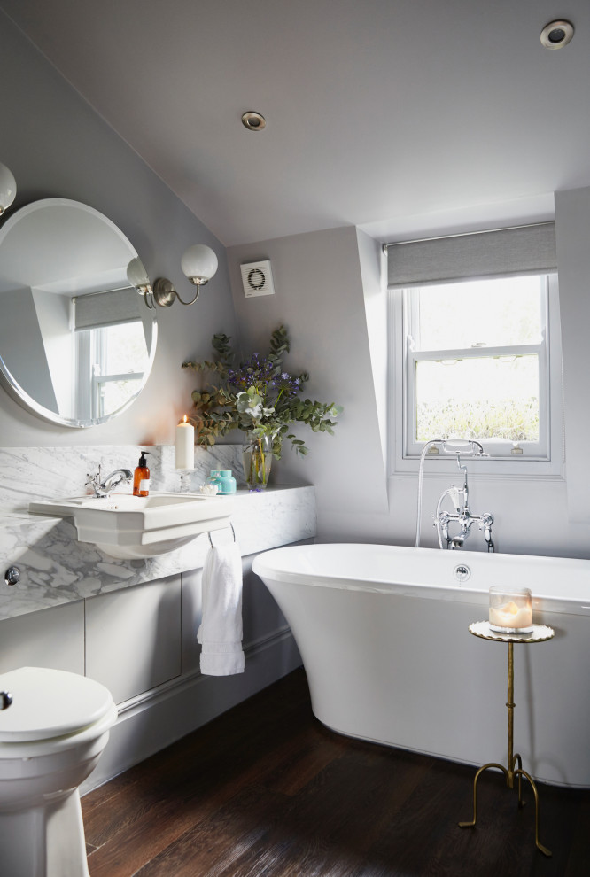 Foto på ett vintage vit badrum, med släta luckor, vita skåp, ett fristående badkar, en toalettstol med hel cisternkåpa, vita väggar, mörkt trägolv, ett fristående handfat och brunt golv