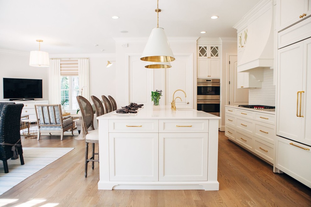 Inspiration for a transitional l-shaped open plan kitchen in Charlotte with a farmhouse sink, recessed-panel cabinets, white cabinets, white splashback, panelled appliances, medium hardwood floors, with island, brown floor and white benchtop.