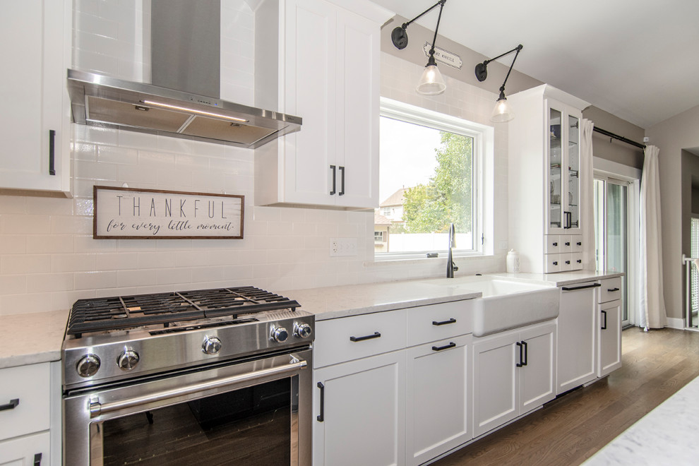 This is an example of an expansive transitional l-shaped eat-in kitchen in Chicago with a farmhouse sink, glass-front cabinets, white cabinets, stainless steel appliances, medium hardwood floors, with island, brown floor, white benchtop and vaulted.